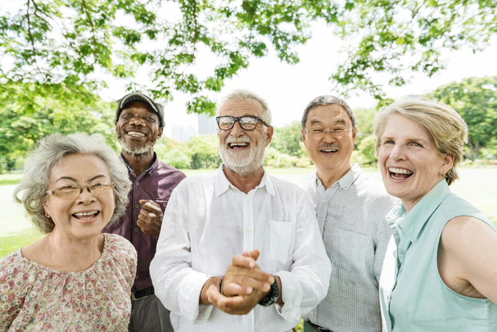 Tiefenbacher Group - A group of five older adults, three men and two women, stand close together outdoors, smiling and laughing under leafy trees on a sunny day, reflecting the joy that patient-centric drug development brings to thriving communities in a lush park setting.