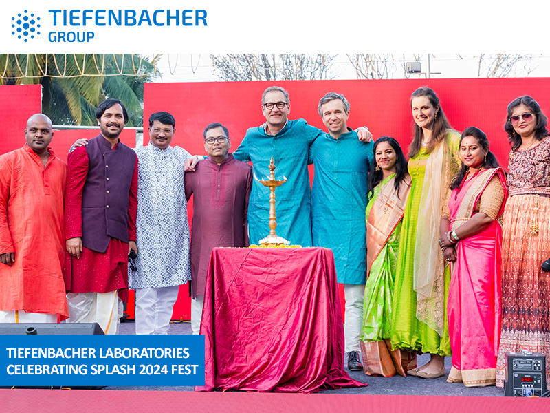 Tiefenbacher Group - A group of people in colorful traditional attire stand smiling on stage around a lit lamp at TIEFENBACHER LABORATORIES SPLASH 2024 Fest, celebrating advancements in pharmaceutical healthcare solutions, with a red backdrop and event signage visible.