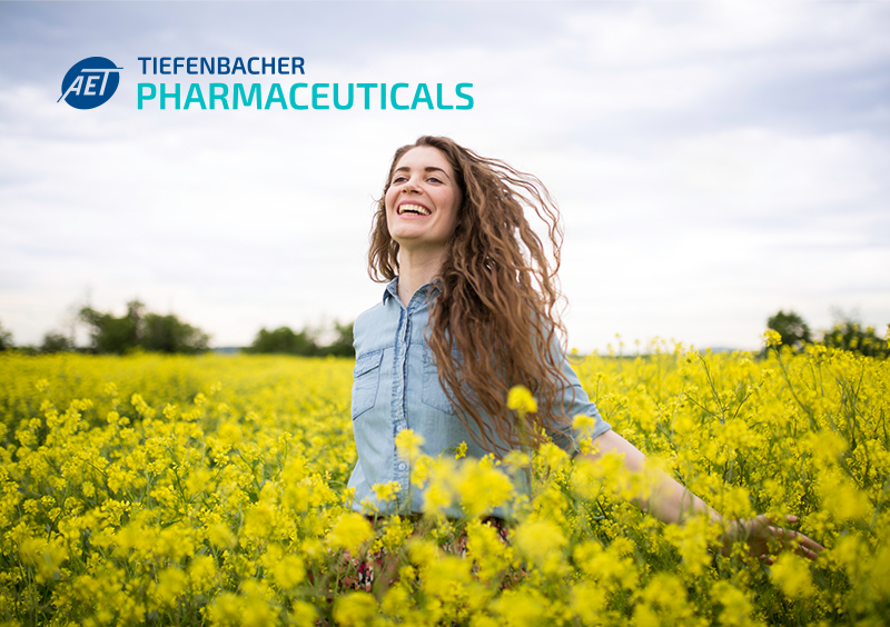 Tiefenbacher Group - A smiling woman with long hair stands in a field of yellow flowers under a clear sky. The Tiefenbacher Pharmaceuticals logo in the top left highlights their dedication to patient-centric drug development and innovative pharmaceutical healthcare solutions.