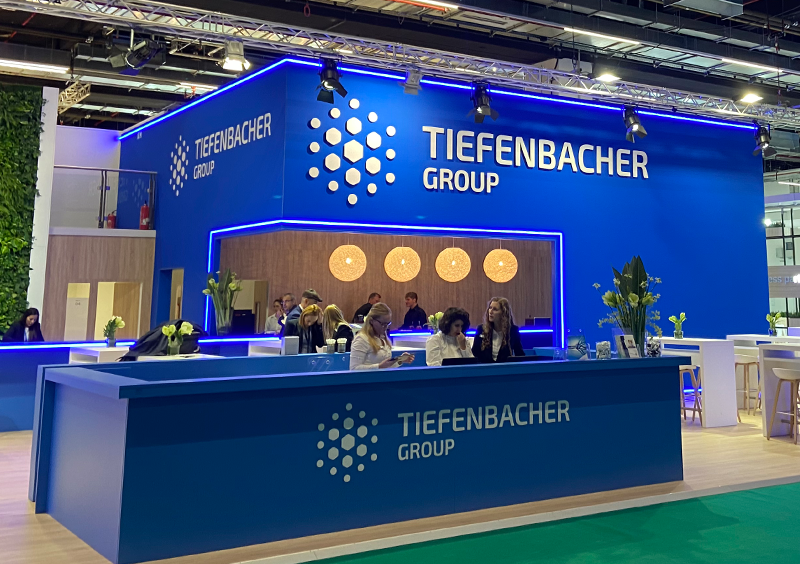 Tiefenbacher Group - A modern exhibition booth for TIEFENBACHER GROUP, a family-owned pharmaceutical company, features a bold blue design, illuminated logo, reception counter, and several people interacting inside a convention or trade show setting.
