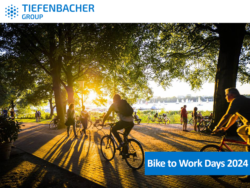 Tiefenbacher Group - People riding bicycles along a sunlit park path near water, with trees providing shade. Overlay text reads "Bike to Work Days 2024." "Tiefenbacher Group," dedicated to pharmaceutical healthcare solutions, is displayed at the top.