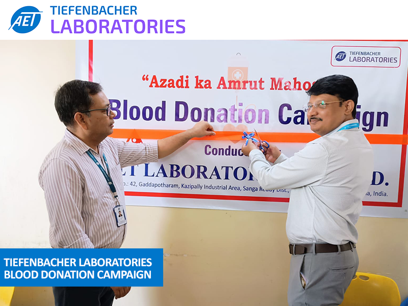 Tiefenbacher Group - Two men in formal attire cut a red ribbon at an event in front of a banner reading "Blood Donation Campaign" at Tiefenbacher Laboratories, a leader in generic medicines manufacturing. A blue label highlights the blood donation campaign.