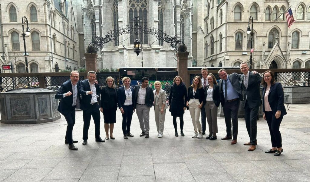 Tiefenbacher Group - A group of thirteen people in business attire stand smiling together outside a historic Gothic-style building, representing leaders in pharmaceutical healthcare solutions and patient-centric drug development.