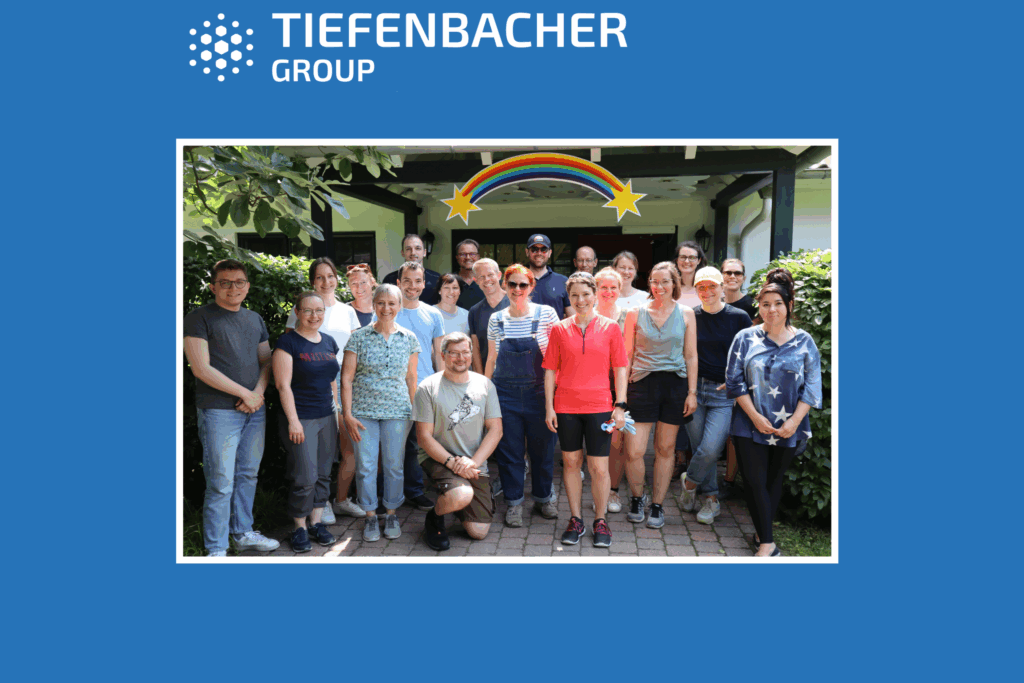 Tiefenbacher Group - A group of about 20 people poses and smiles outdoors under a rainbow decoration, with trees and a building in the background. The blue border features the "Tiefenbacher Group" logo, highlighting their focus on patient-centric drug development.