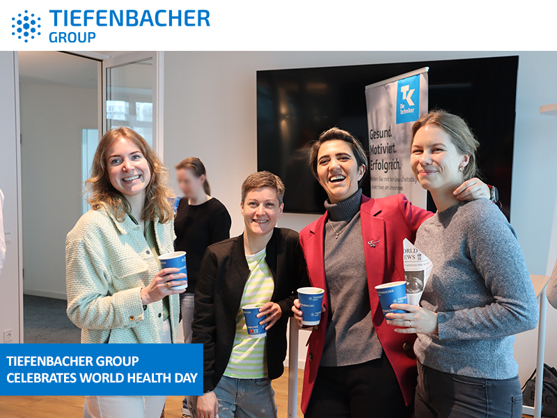 Tiefenbacher Group - Four people stand together indoors, smiling and holding cups. A banner and TV screen highlight the Tiefenbacher Group, a family-owned pharmaceutical company, celebrating World Health Day.