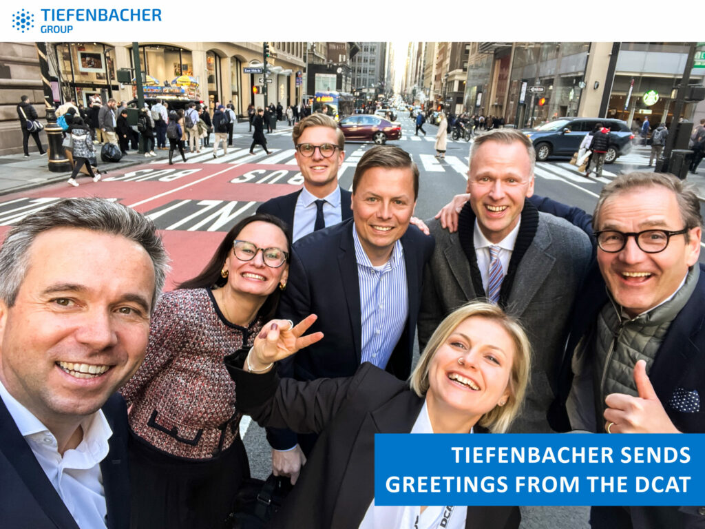 Tiefenbacher Group - A group of seven smiling people in business attire take a selfie on a busy city street. A banner reads, "Tiefenbacher sends greetings from the DCAT." The TIEFENBACHER GROUP logo highlights this family-owned pharmaceutical company.