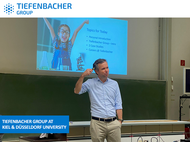 Tiefenbacher Group - A man speaks in front of a chalkboard and a projected slide listing discussion topics. The Tiefenbacher Group logo and the text "Tiefenbacher Group at Kiel & Düsseldorf University" highlight its role in generic medicines manufacturing.