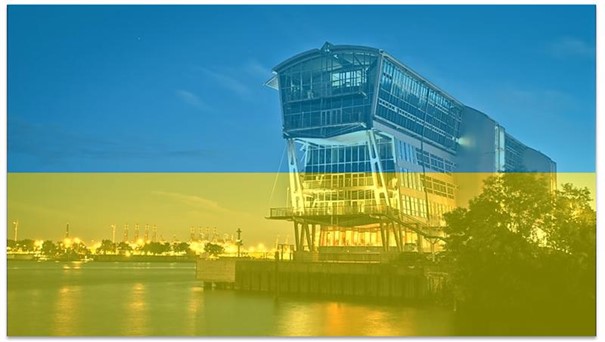 Tiefenbacher Group - A modern riverside building at dusk with city lights in the background, overlaid with a transparent blue and yellow filter resembling the Ukrainian flag, symbolizing innovative pharmaceutical healthcare solutions.