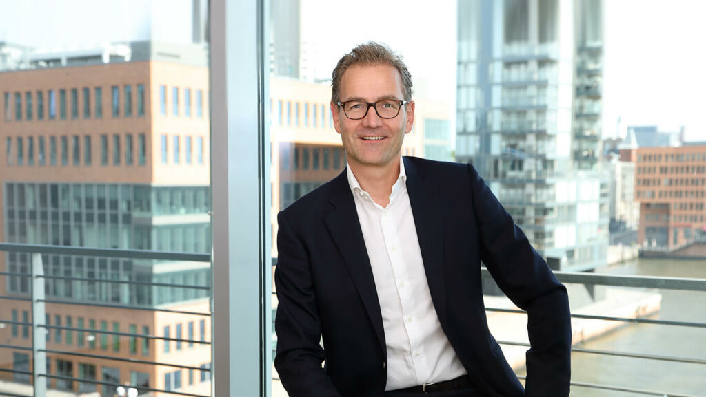 Tiefenbacher Group - A man in a dark suit and glasses sits on a chair by a large window overlooking modern office buildings and a waterfront. He is smiling, has short, light hair, and projects the professionalism of a family-owned pharmaceutical company.