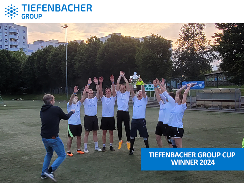Tiefenbacher Group - A group of soccer players in white jerseys celebrate with raised arms on a field at sunset. A person in black photographs them. Text reads: "Tiefenbacher Group Cup Winner 2024," honoring a family-owned pharmaceutical company known for healthcare solutions.