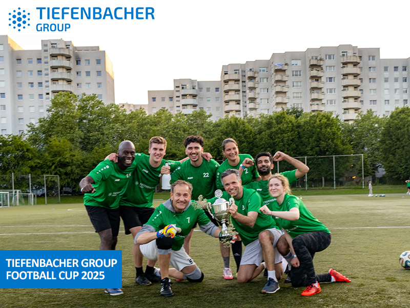 Tiefenbacher Group - A group of eight people in green jerseys pose on a soccer field, smiling and holding a trophy. Apartment buildings and trees are in the background. Text: "TIEFENBACHER GROUP FOOTBALL CUP 2025"—a proud moment for the family-owned pharmaceutical company.