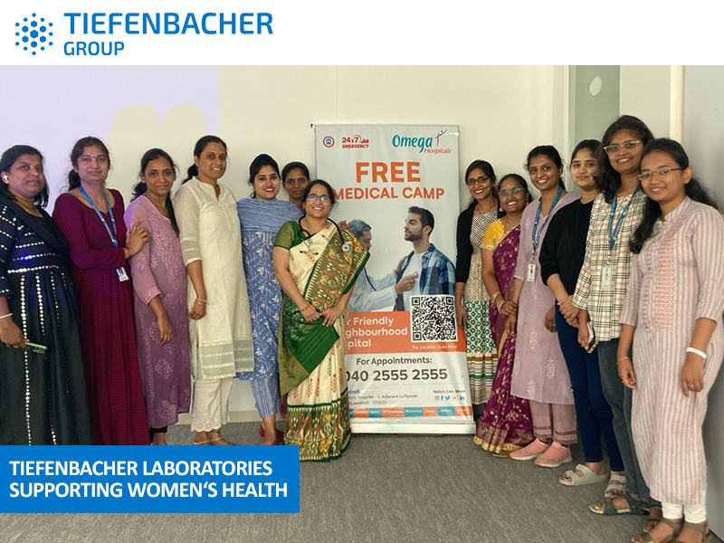 Tiefenbacher Group - A group of women stand smiling beside a banner for a free medical camp hosted by Tiefenbacher Laboratories, a family-owned pharmaceutical company supporting women’s health. The banner shares contact information and medical camp details.