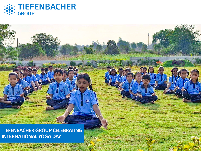 Tiefenbacher Group - A group of schoolchildren sits cross-legged on grass, practicing yoga outdoors with trees in the background. A banner reads, “Tiefenbacher Group, a family-owned pharmaceutical company, celebrating International Yoga Day.”.