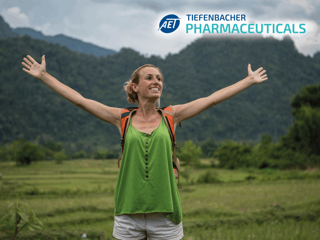 Tiefenbacher Group - A smiling woman in a green top and backpack stands with arms outstretched before lush mountains and fields. The logo and text "Tiefenbacher Pharmaceuticals" highlights their pharmaceutical healthcare solutions in the upper right corner.