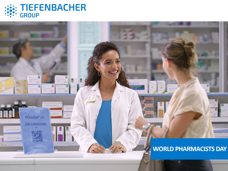 Tiefenbacher Group - A smiling pharmacist in a white coat talks to a customer at the pharmacy counter. Shelves with medicine are in the background, highlighting pharmaceutical healthcare solutions. Text reads "World Pharmacists Day.