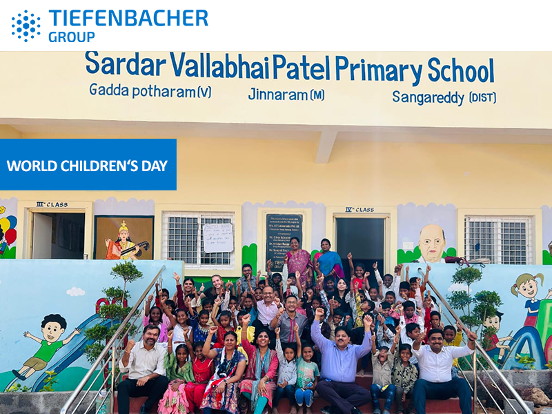 Tiefenbacher Group - A large group of children, teachers, and adults pose and smile outside Sardar Vallabhbhai Patel Primary School, decorated with colorful murals. A banner reads “World Children’s Day,” celebrating advancements like patient-centric drug development.