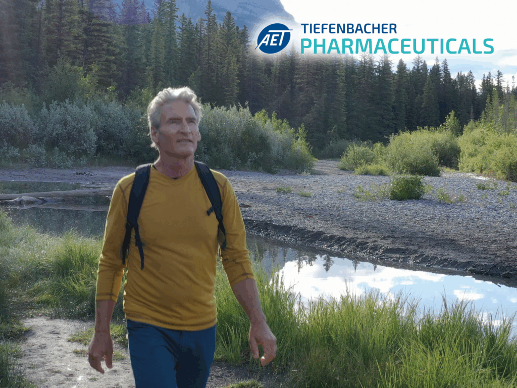 Tiefenbacher Group - An older man with gray hair, wearing a yellow long-sleeve shirt and backpack, walks in a lush riverside landscape. In the corner, the logo and text read "AET TIEFENBACHER PHARMACEUTICALS," a family-owned pharmaceutical company.