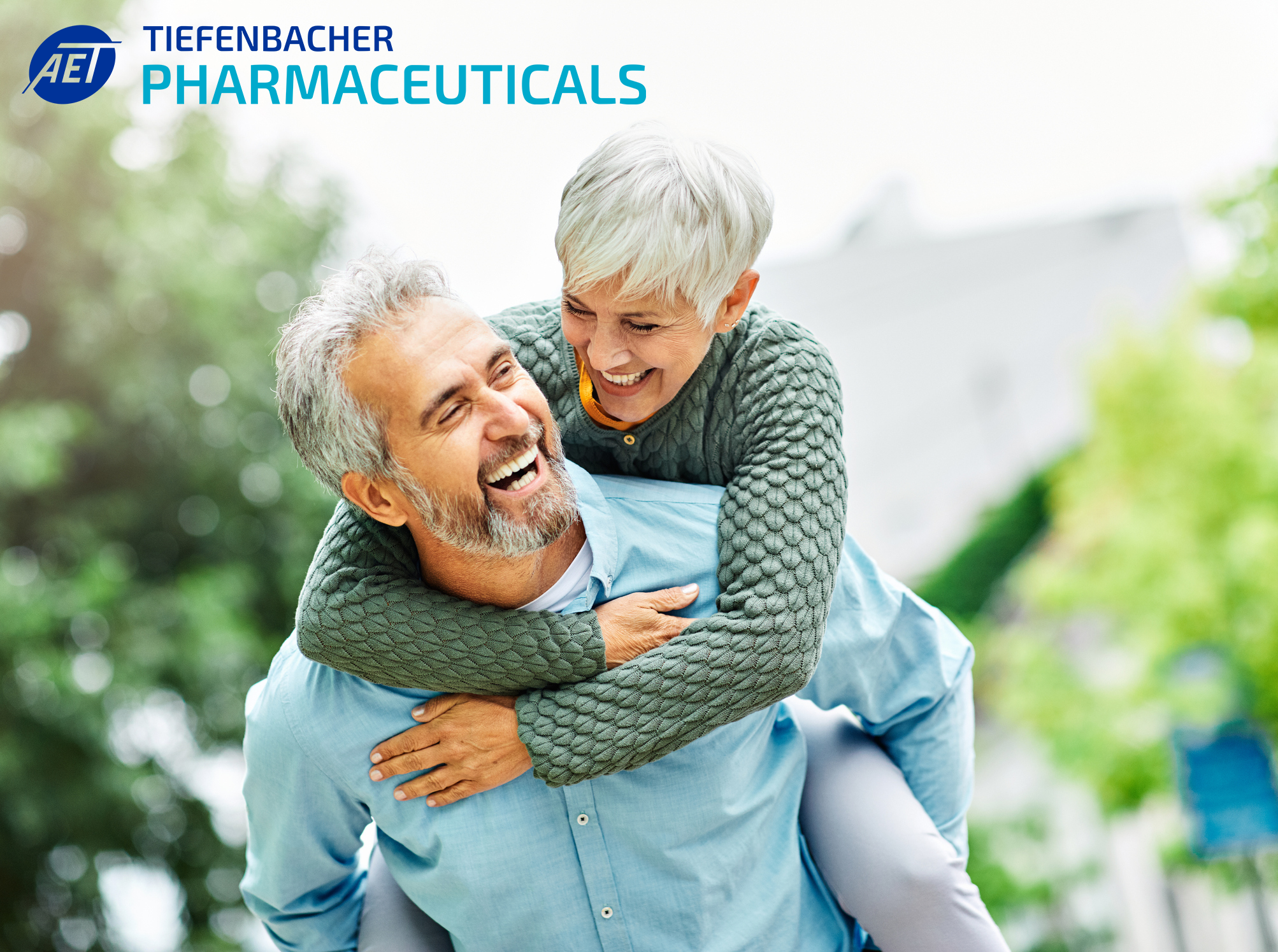 Tiefenbacher Group - A smiling older woman with short gray hair joyfully gives a piggyback ride to a laughing older man outdoors, highlighting Tiefenbacher Pharmaceuticals' commitment to generic medicines manufacturing. The company logo is in the top left corner.